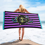 Purple and black striped towel with teal detail and snake and banter print, being held up in the corners horizontally on a beach
