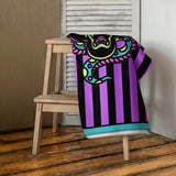 Purple and black striped towel with teal detail and snake and banter print, folded and hanging from a towel stand inside a house